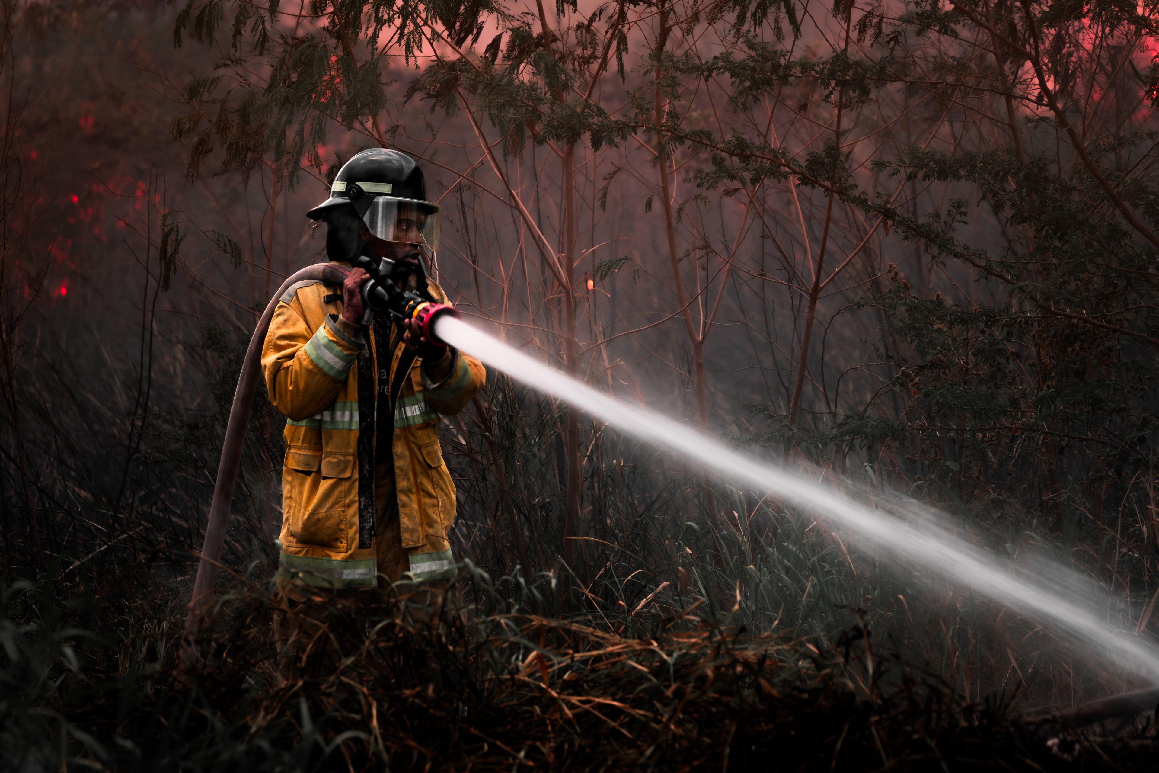 A firefighter in a yellow turnout coat and visored helmet sprays a jet of water into smoky bushland glowing red with fire.