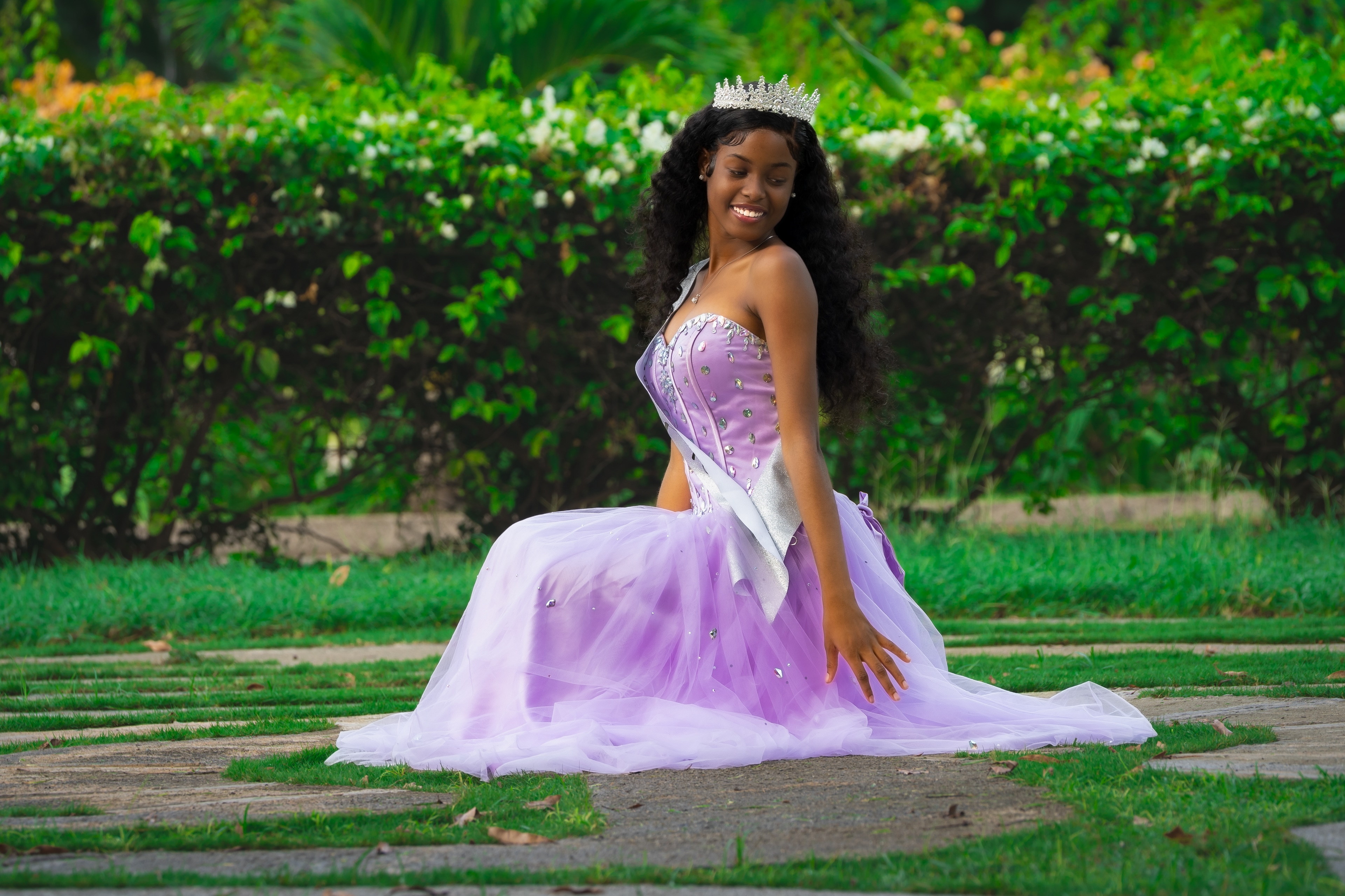 A smiling girl in a crystal tiara, sash and lilac tulle gown sits on a grass-and-stone path, looking down to her shoulder before a flowering hedge.