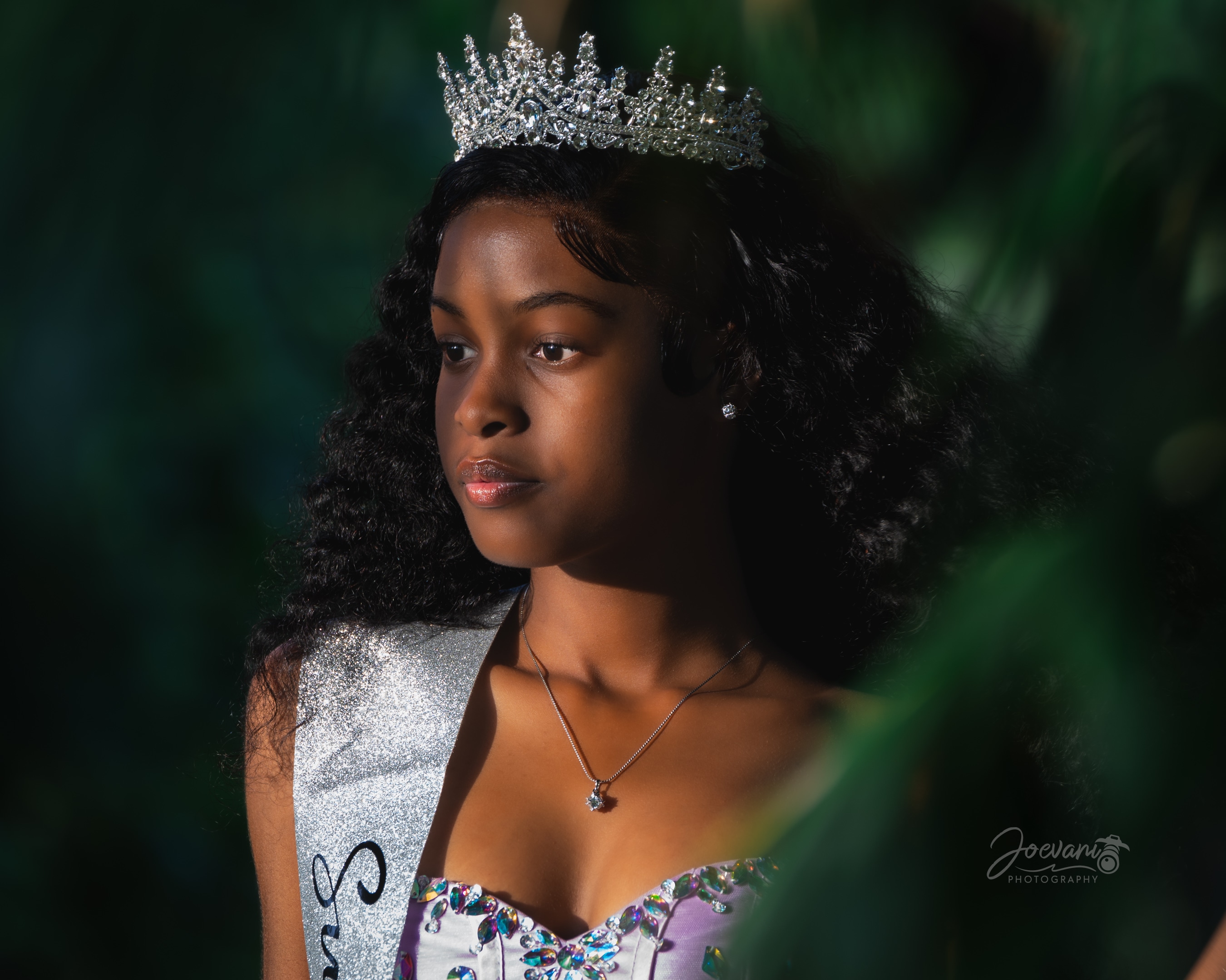 A girl in a crystal tiara, silver sash and jewelled lilac bodice gazes aside in close-up, framed by dark leaves in warm low sunlight.