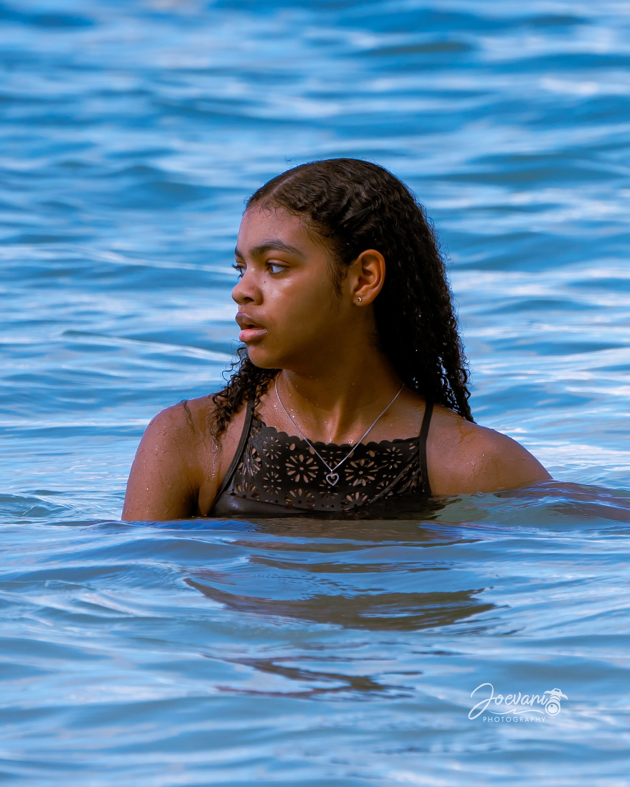 A girl with long wet curls and a heart pendant stands shoulder-deep in rippling blue sea, looking off to one side in bright sunlight.