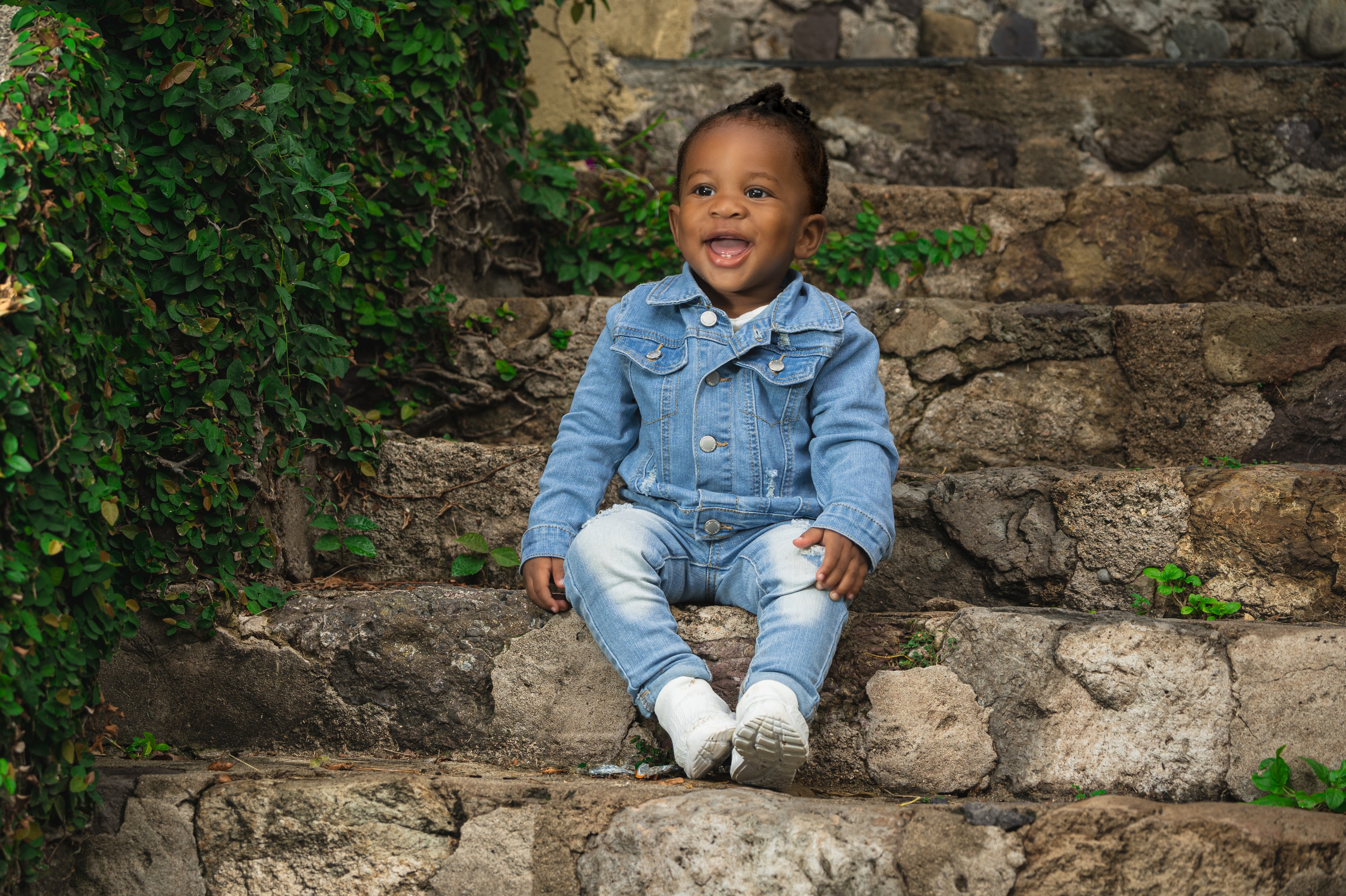 A laughing toddler in a denim jacket, light distressed jeans and white shoes sits on old stone steps beside trailing green vines.