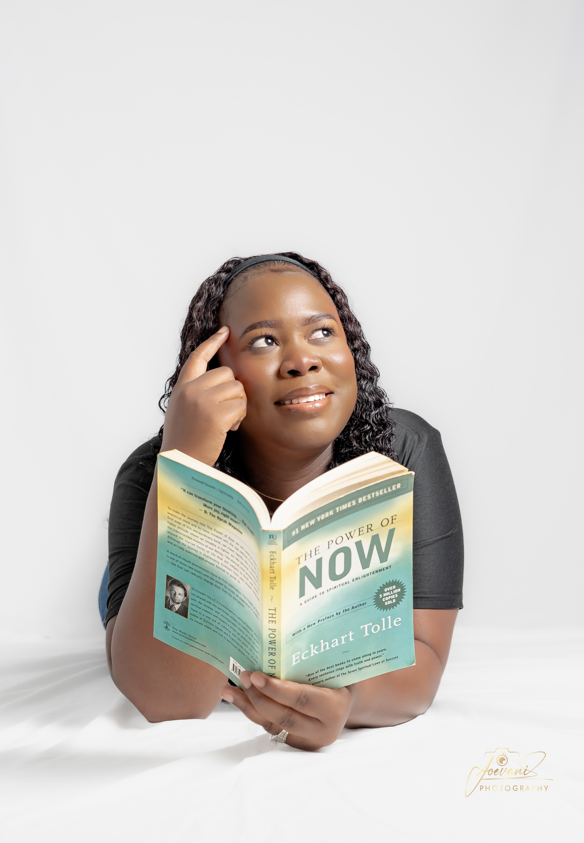 A smiling woman lies on her front holding an open copy of The Power of Now, finger to temple and gazing upward, on a white studio backdrop.