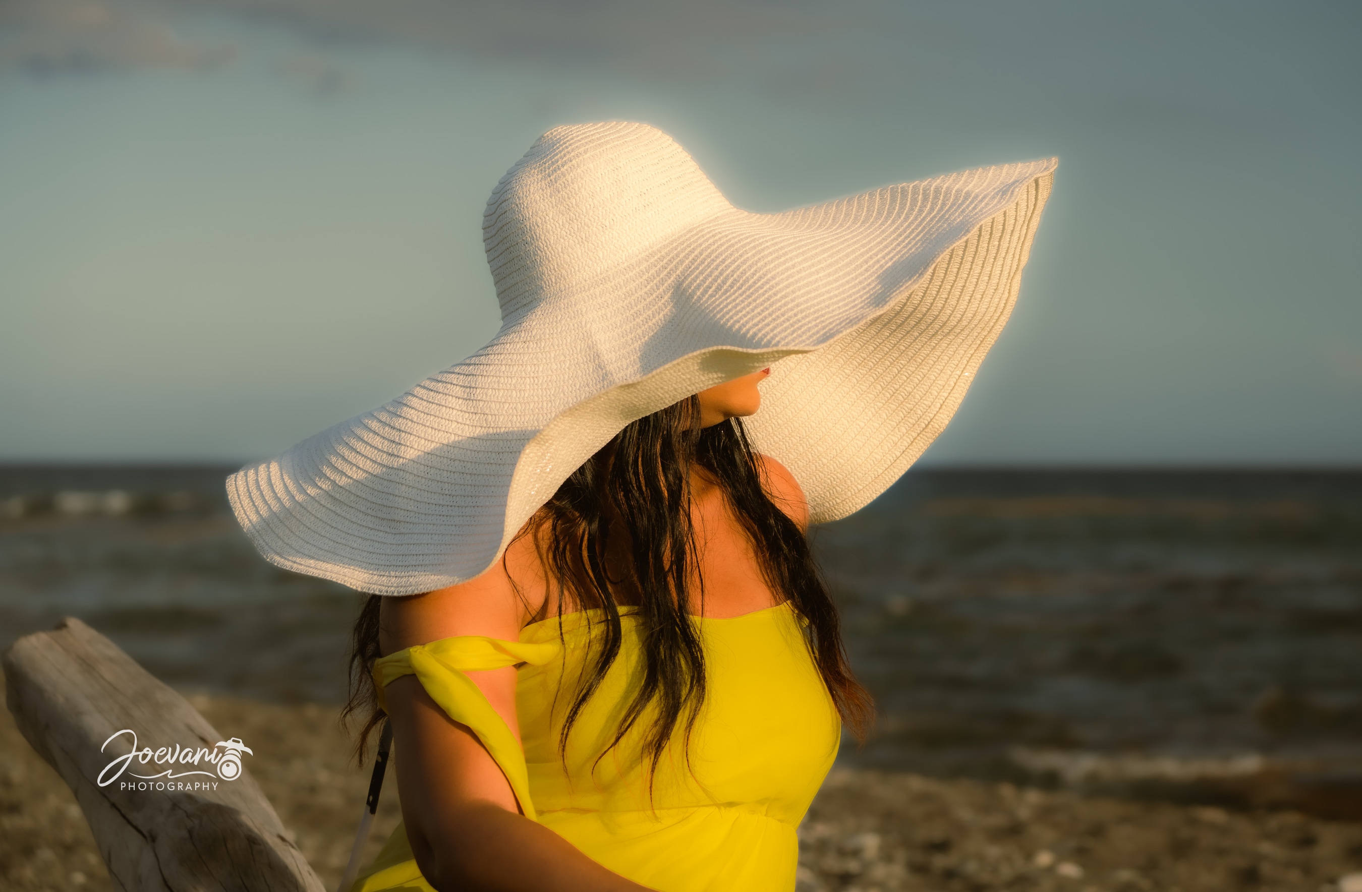 A woman in a yellow off-shoulder dress turns away beneath an oversized white sun hat beside driftwood by the sea in low golden light.