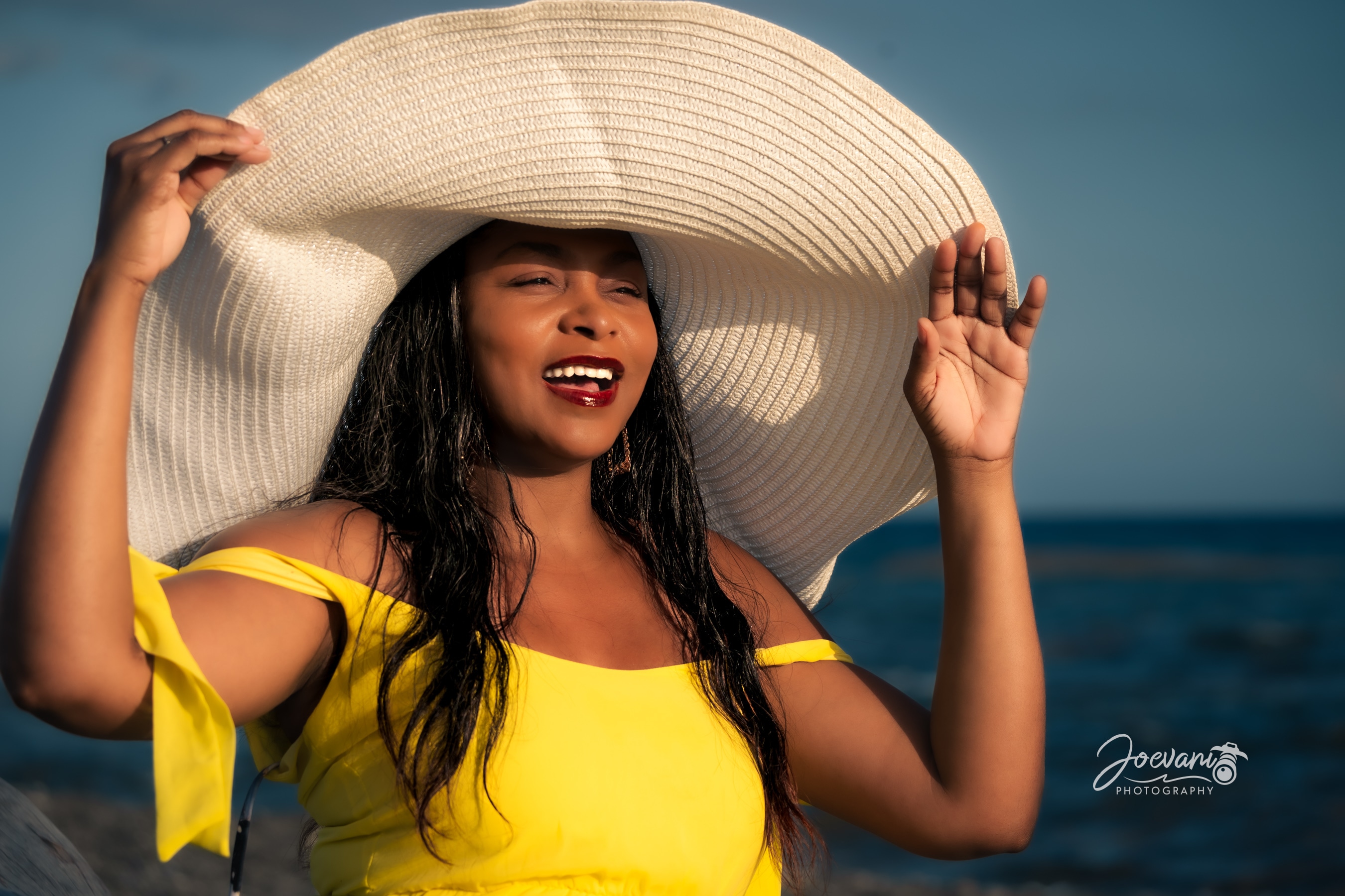 A laughing woman in a yellow off-shoulder dress holds the brim of an oversized white sun hat with one hand, the other raised, blue sea behind.